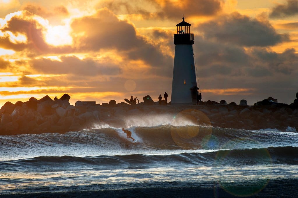 A surfer riding a wave at sunset with the Walton Lighthouse in the background in Santa Cruz, California.