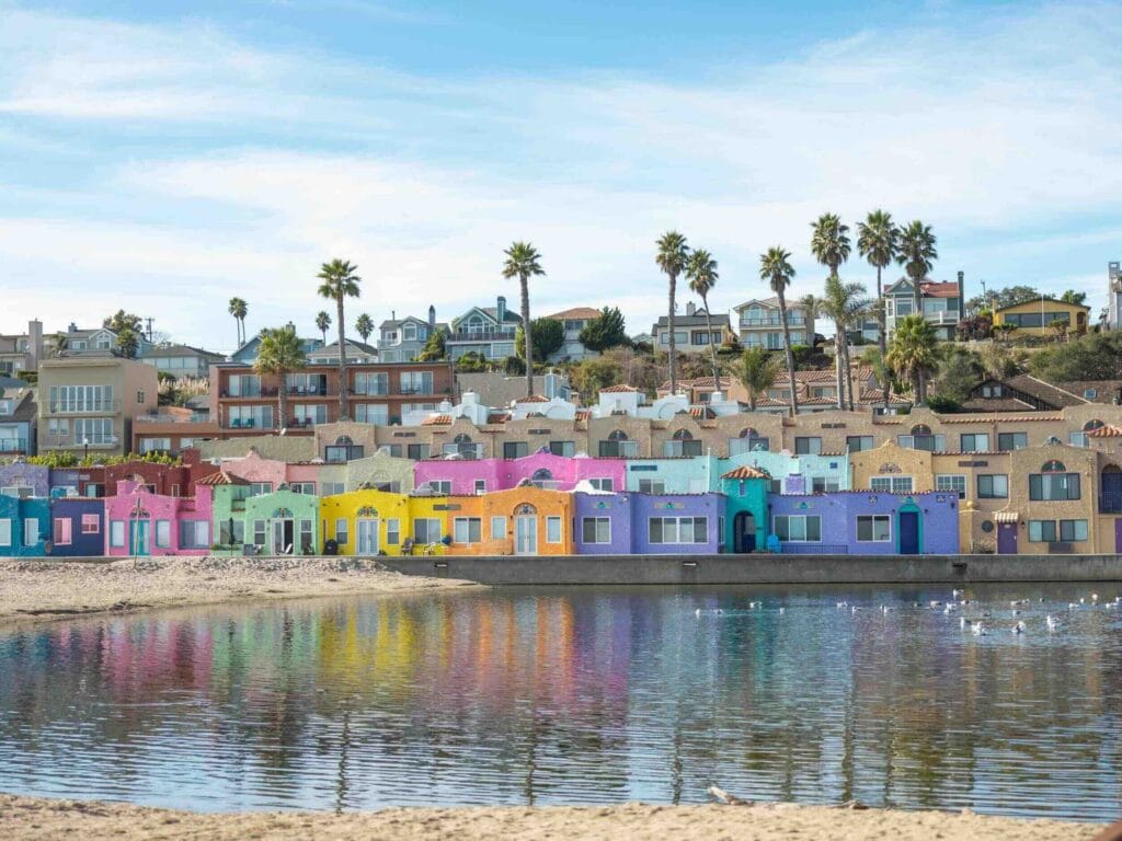 Colorful beachfront houses in Capitola, California, reflecting on the calm waters of the lagoon under a clear blue sky.