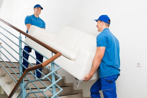 Two professional movers carrying a white sofa up a staircase in a residential building.