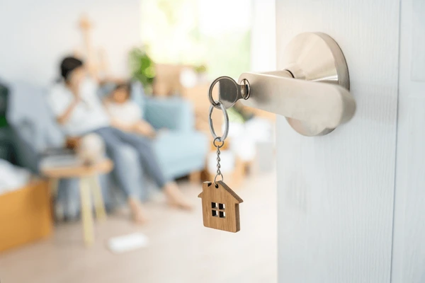 Close-up of a key with a house keychain in a door, with a family sitting on a couch in the background during an apartment move.