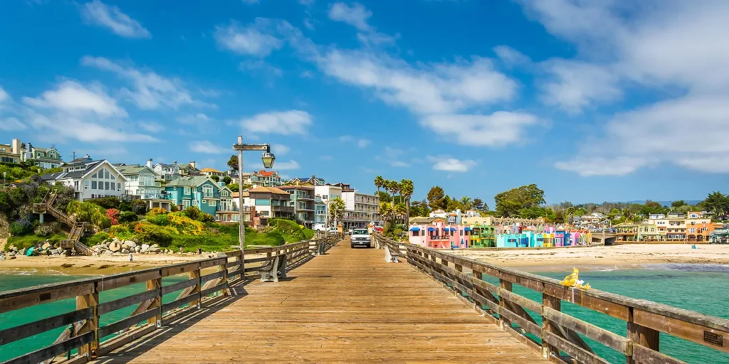 A scenic view of the Capitola Wharf leading to colorful beachfront houses in Capitola, California, under a bright blue sky.