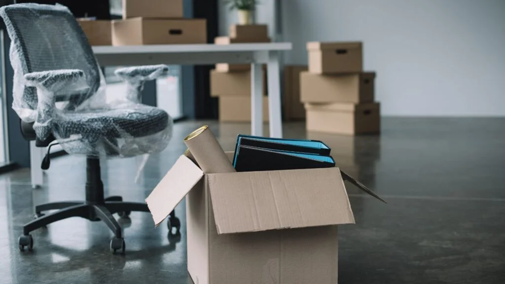 A wrapped office chair and packed boxes, including a cardboard box with folders and supplies, in an office space.